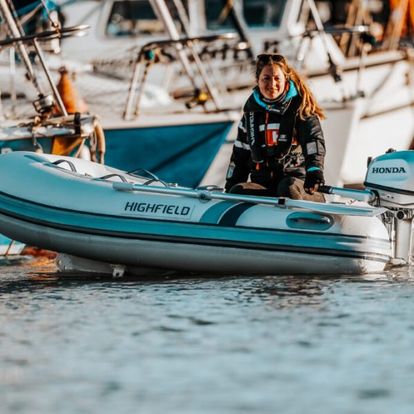 Mujer navegando en una lancha inflable Highfield con motor Honda, en un puerto con barcos de fondo. La mujer lleva un chaleco salvavidas y sonríe mientras se encuentra en el agua.