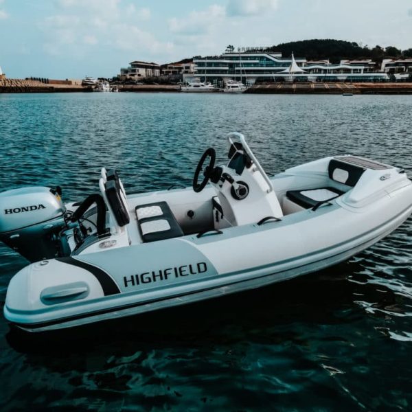 Lancha inflable Highfield con motor Honda en aguas tranquilas, frente a un puerto moderno y un faro. Ideal para actividades acuáticas y paseos en el mar.
