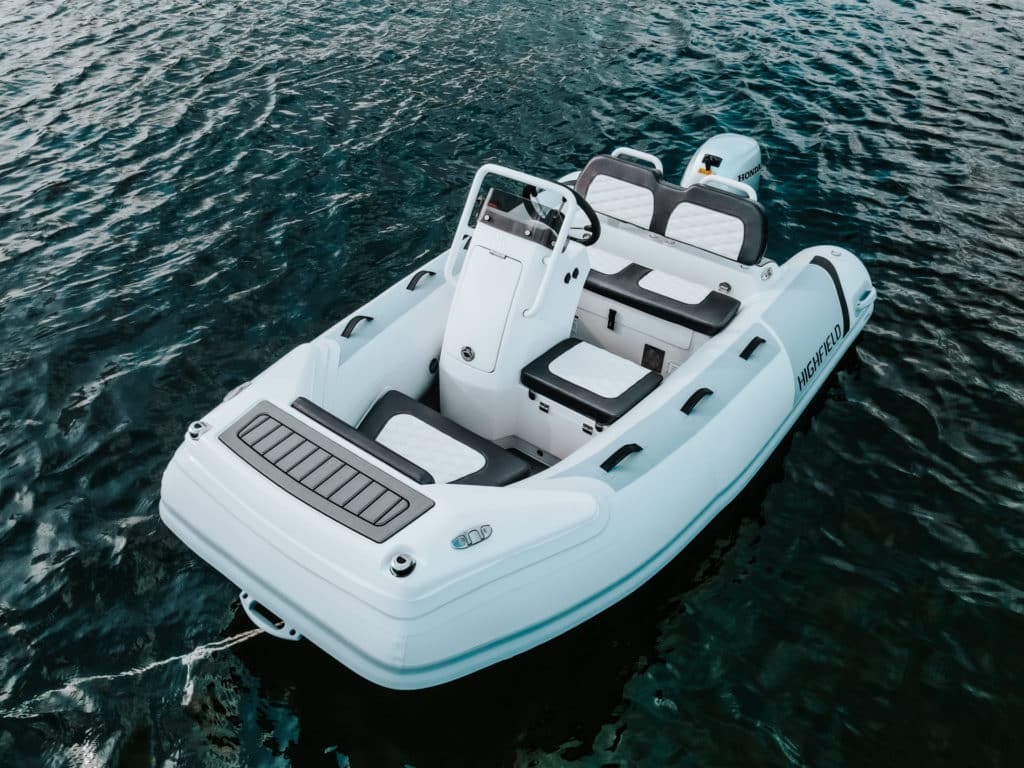 Bote inflable Highfield blanco sobre aguas tranquilas, con asientos en la parte interior y motor Honda en la parte trasera. Ideal para actividades acuáticas y excursiones.