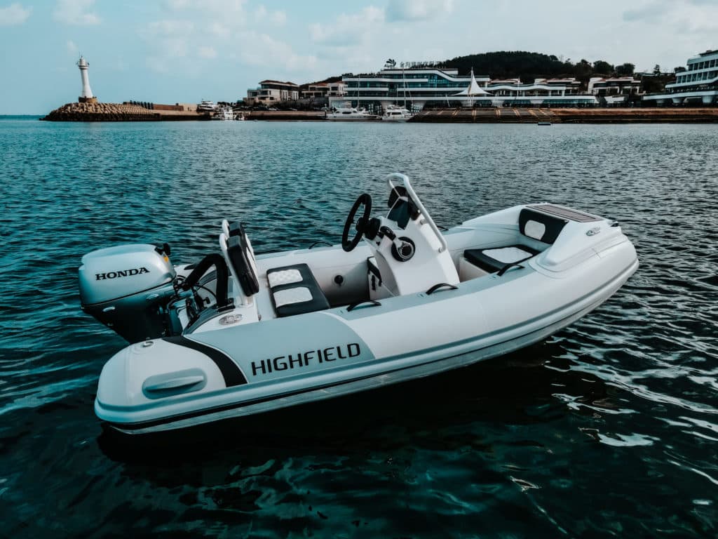 Lancha inflable Highfield con motor Honda en aguas tranquilas, frente a un puerto moderno y un faro. Ideal para actividades acuáticas y paseos en el mar.