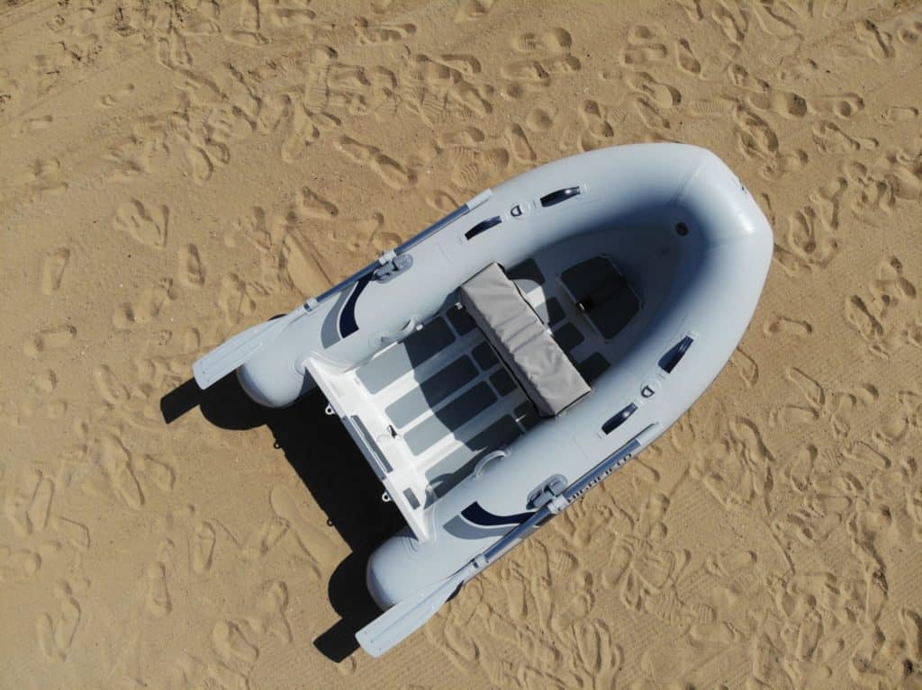 Bote inflable gris sobre arena, visto desde arriba, con un asiento central y espacio para varios ocupantes, ideal para actividades acuáticas y exploración.